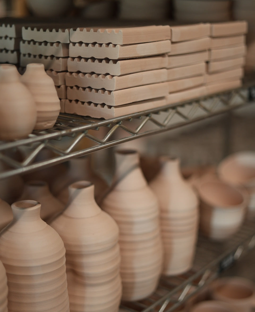 Unglazed ceramic pottery pieces in a natural light tan clay color, including bottles, bowls, and stacked rectangular tiles, displayed on metal shelving.