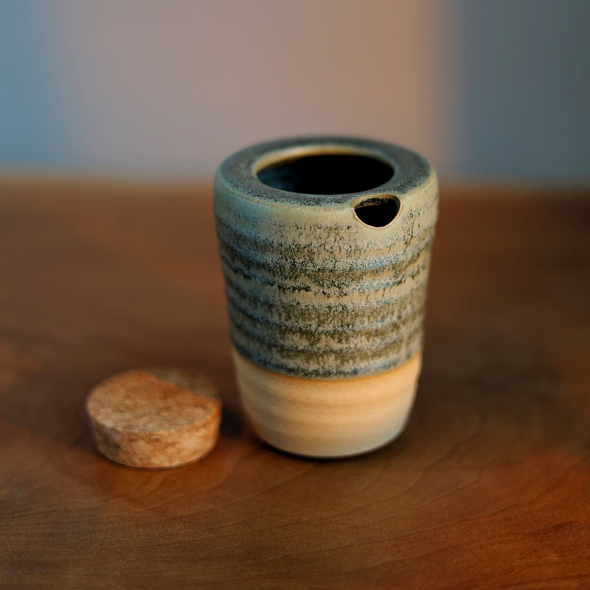 A small handmade ceramic vessel with a textured blue and beige glaze sits on a wooden surface next to a cork stopper, with a softly blurred blue background.