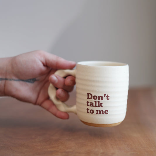 Hand holding a cream handmade ceramic mug with 'Don't talk to me' text on a wooden surface.