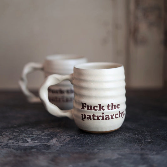 White ceramic mug with 'Fuck the patriarchy' text on a dark surface.