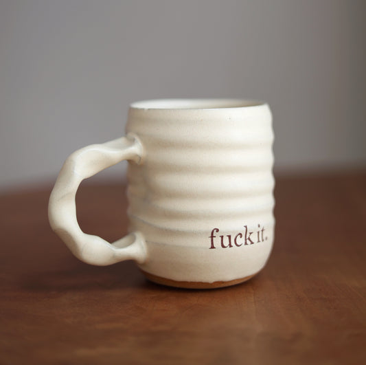 Off White ceramic mug with 'fuck it.' text on a wooden surface and gray background