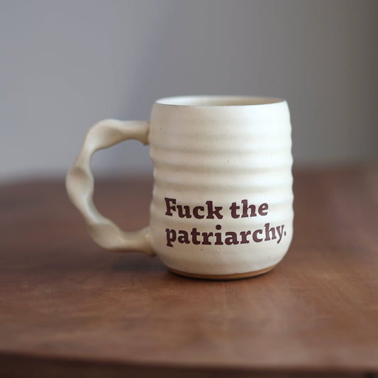 White mug with 'Fuck the patriarchy.' text on a wooden surface