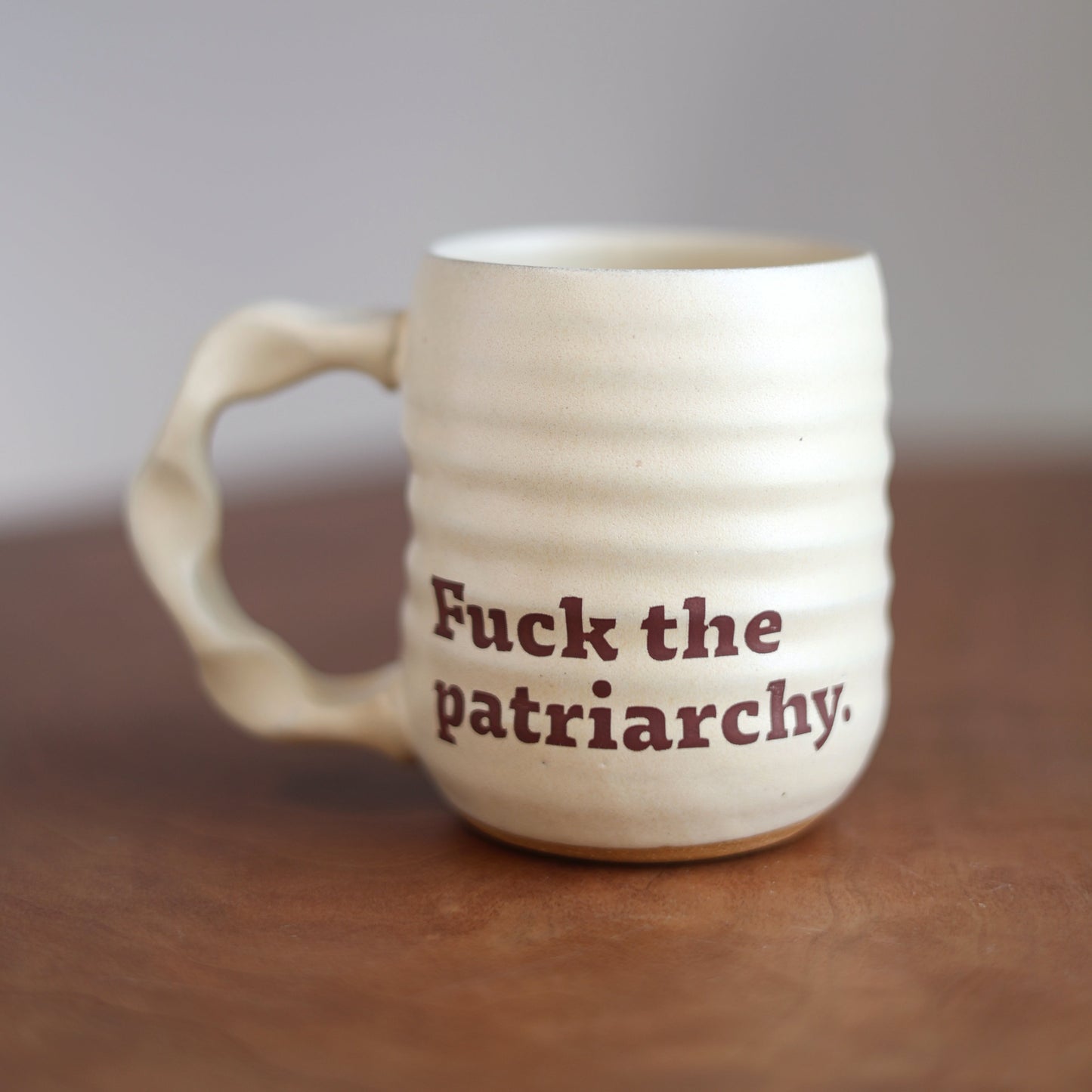 White ceramic mug with 'Fuck the patriarchy.' text on a wooden surface.