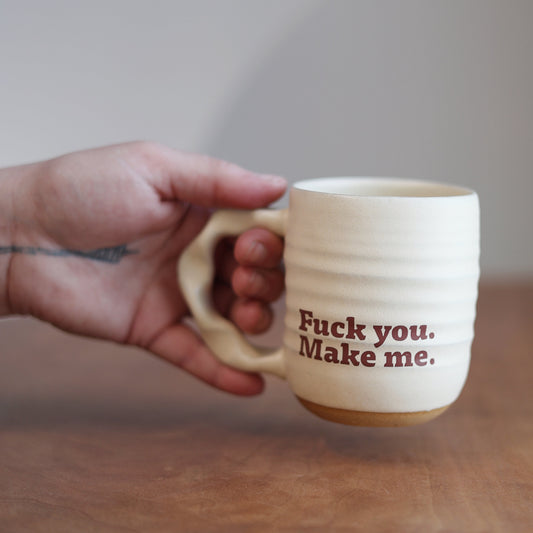 Hand holding a mug with text “fuck you make me” on a wooden surface