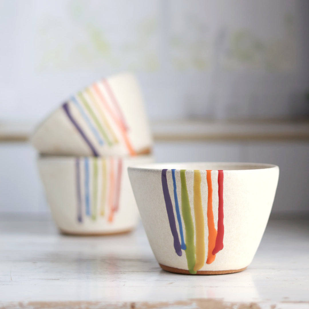 "Stack of three cream colored ceramic bowls with rainbow drip glaze patterns, artistically arranged showing the colorful striped designs"