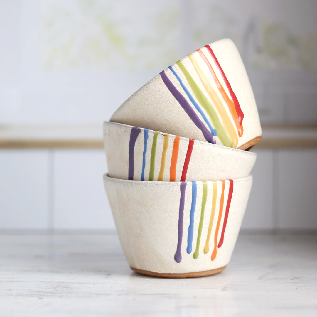 "Stack of three cream colored ceramic bowls with rainbow drip glaze patterns, artistically arranged showing the colorful striped designs"