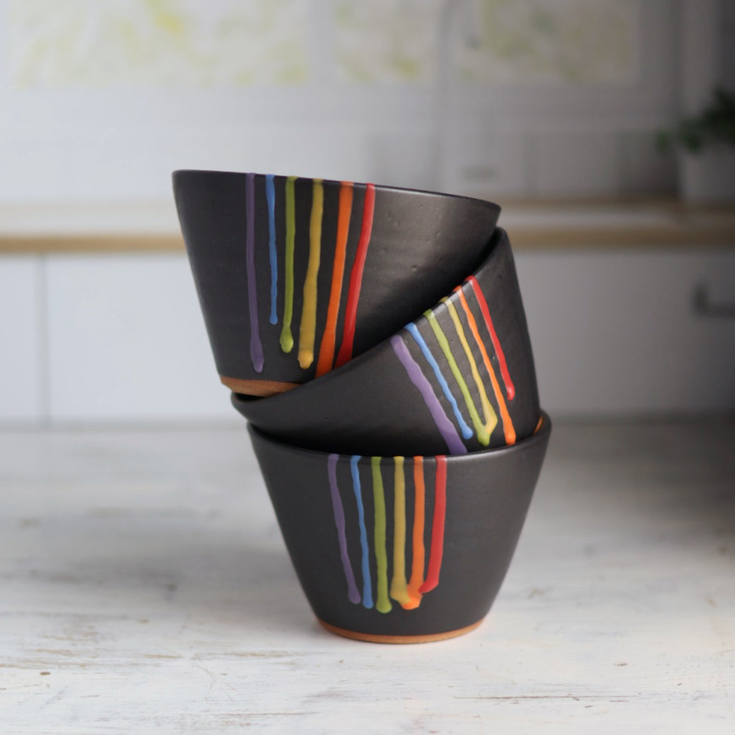 "Three black ceramic bowls stacked at angles showing rainbow drip glaze patterns with vibrant colored stripes against dark background"