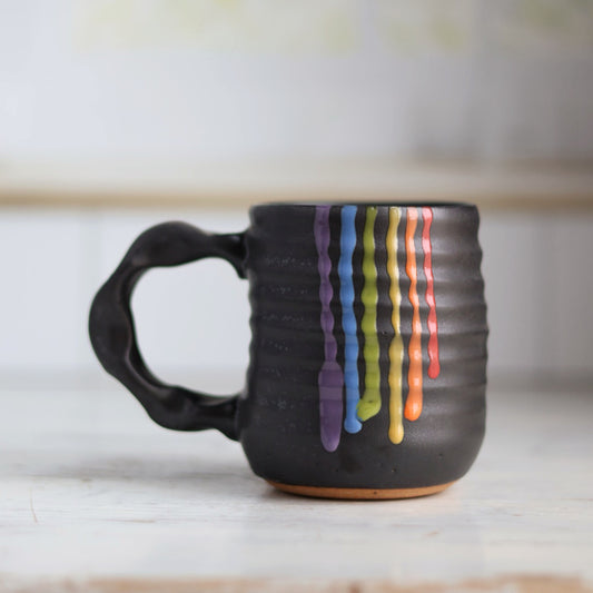 "Black ceramic companion mug with vibrant rainbow colored glaze drips cascading down the side, showing purple, blue, green, yellow, orange and red stripes"