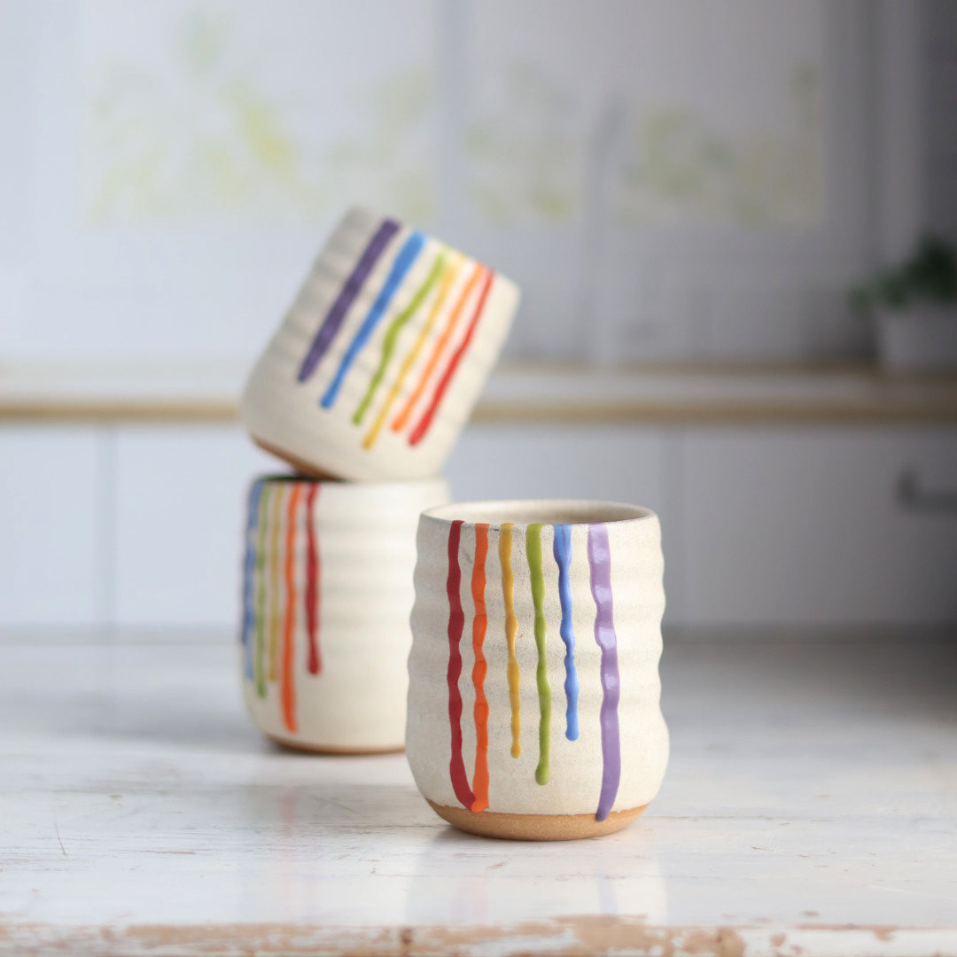 "Cream colored ceramic wine cup featuring rainbow drip glaze pattern with red, orange, yellow, green, blue and purple flowing down the sides"