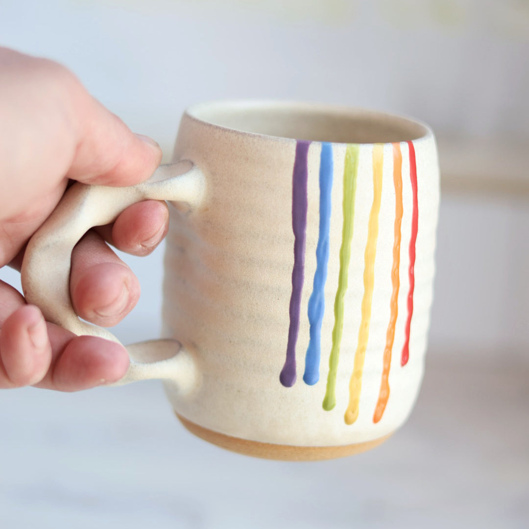 "Cream colored ceramic companion mug with handle featuring rainbow drip glaze pattern with purple, blue, green, yellow, orange and red stripes flowing down the side"