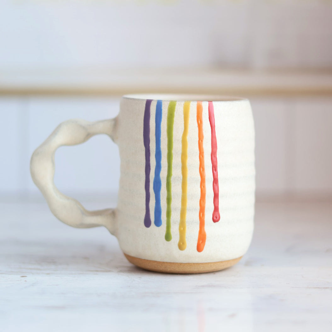 "Cream colored ceramic companion mug with handle featuring rainbow drip glaze pattern with purple, blue, green, yellow, orange and red stripes flowing down the side"