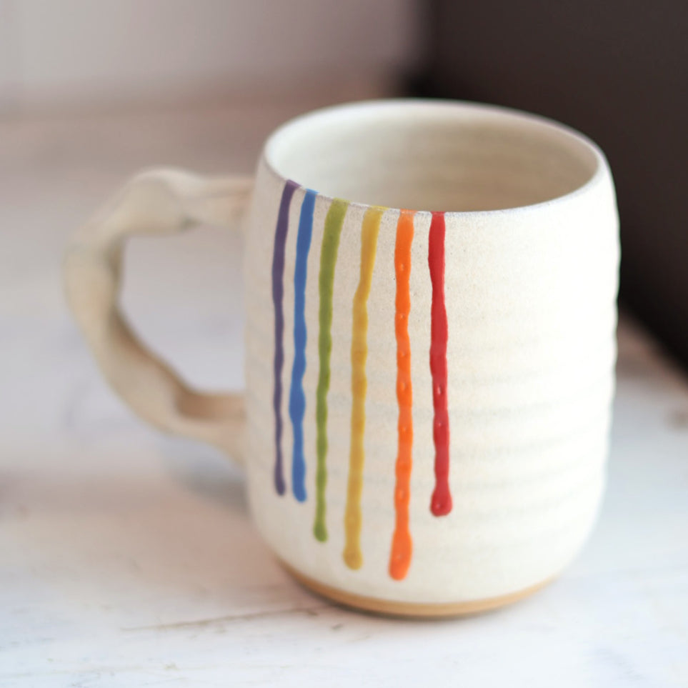 "Cream colored ceramic companion mug with handle featuring rainbow drip glaze pattern with purple, blue, green, yellow, orange and red stripes flowing down the side"
