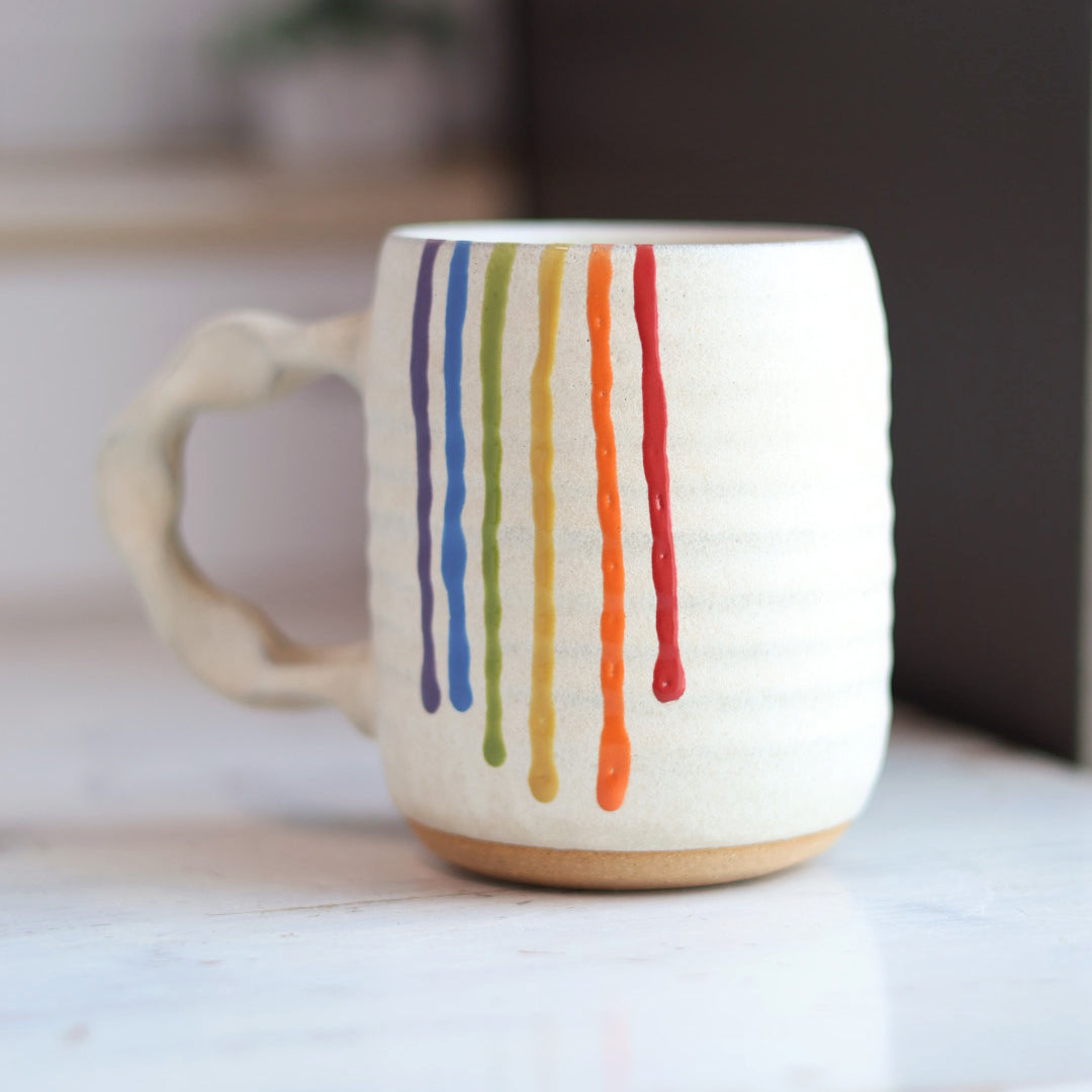 "Cream colored ceramic companion mug with handle featuring rainbow drip glaze pattern with purple, blue, green, yellow, orange and red stripes flowing down the side"