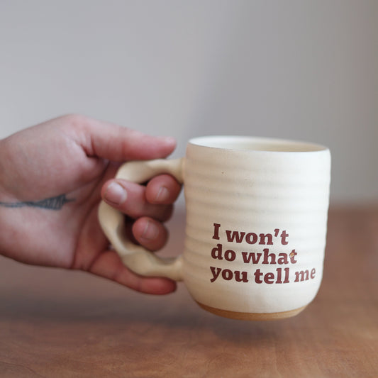 Person holding a mug with text “I won’t do what you tell me” on a wooden surface