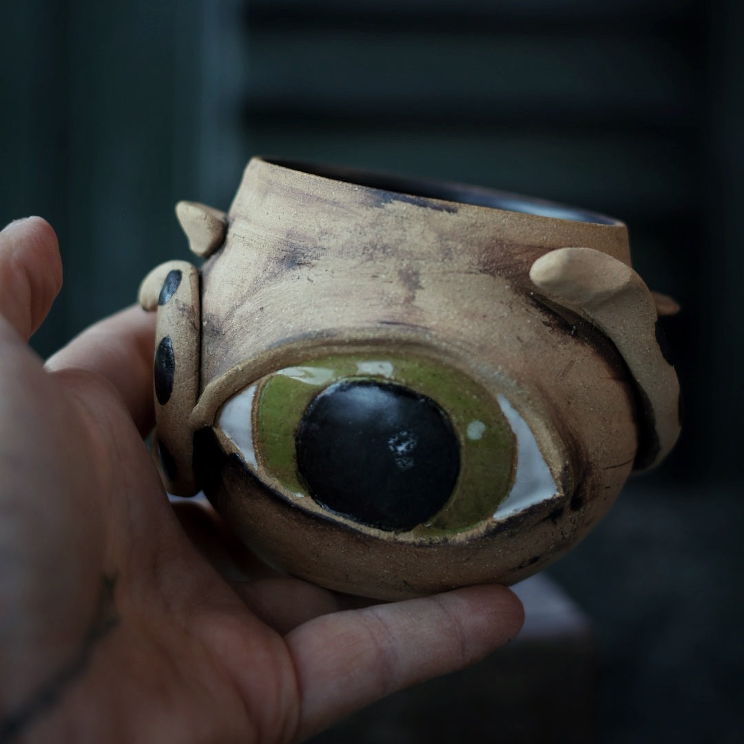Hand holding a ceramic cauldron with one large sculpted eye against a dark background
