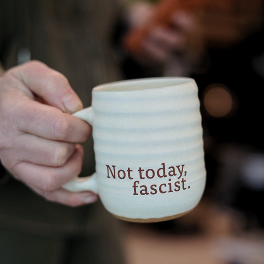 Person holding a mug with 'Not today, fascist.' text in a blurred indoor setting