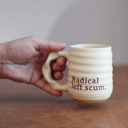 Person holding a mug with 'Radical left scum' text on a wooden table.