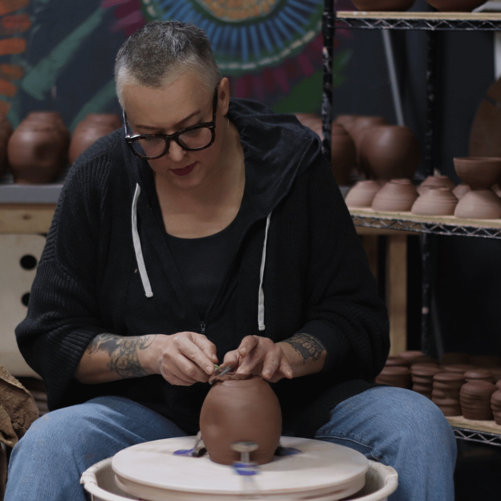 A person with short gray hair and glasses sits at a pottery wheel, shaping a small terracotta pot. Shelves in the background are filled with finished clay vessels and pottery pieces.