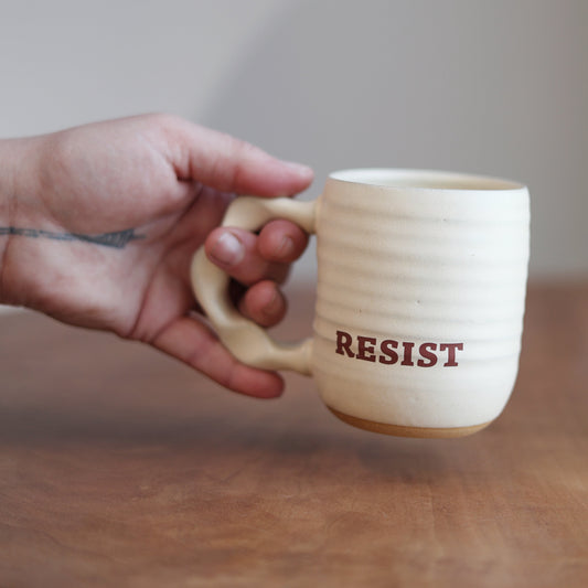 Hand holding a mug with 'RESIST' printed on it against a neutral background