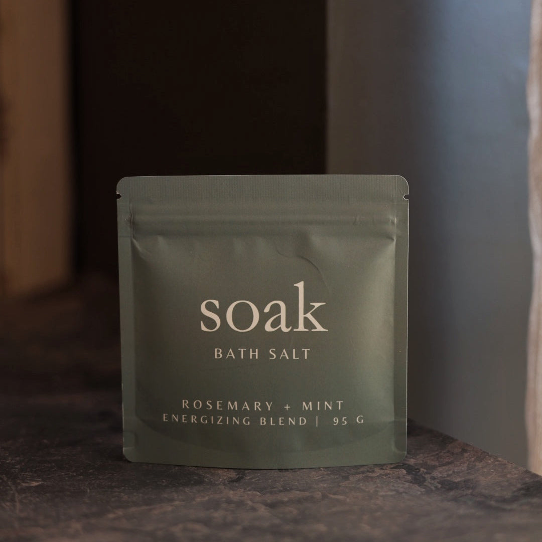 Green package of 'soak' bath salt with 'Rosemary + Mint' flavor on a dark surface.