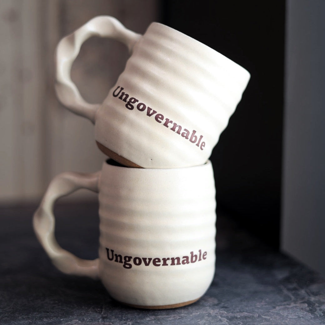 Two white handmade mugs with 'Ungovernable' text stacked on a dark surface.
