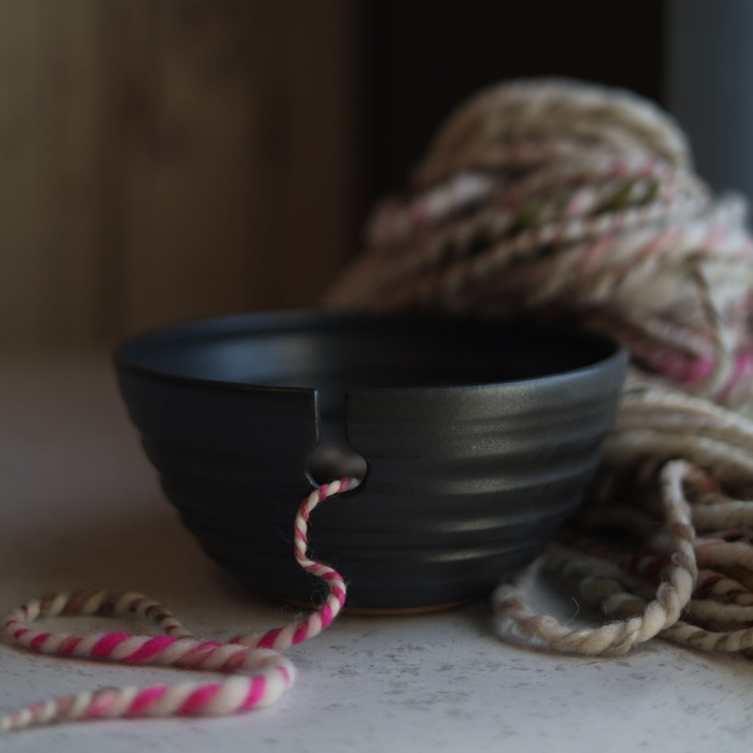 Black ceramic yarn bowl with pink and white striped yarn on a neutral background