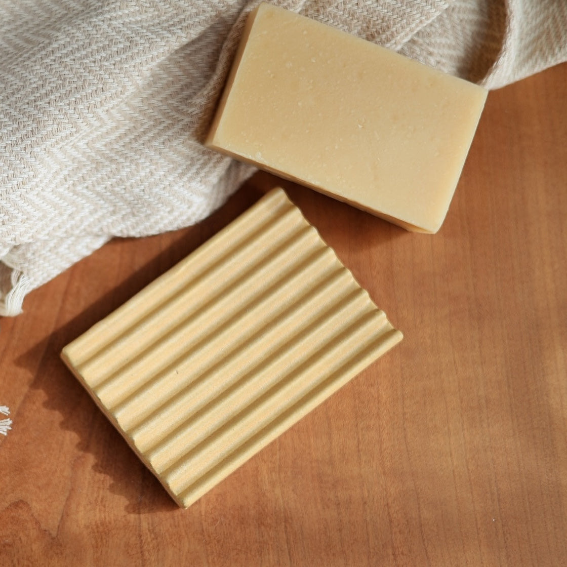One beige soap bar and a ceramic soap dish on a wooden surface with a white cloth in the background