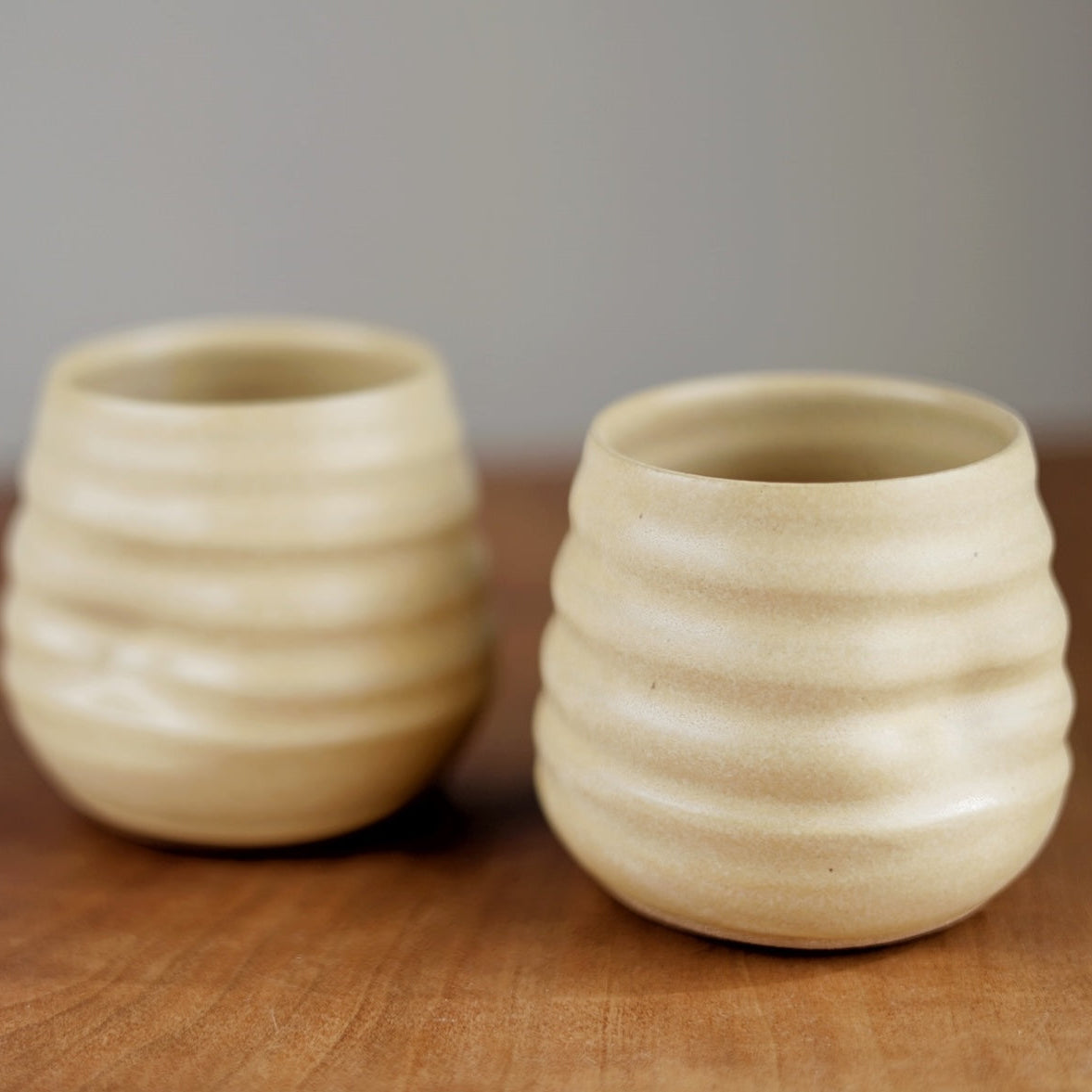 Two beige ceramic cups with textured surface on a wooden surface.