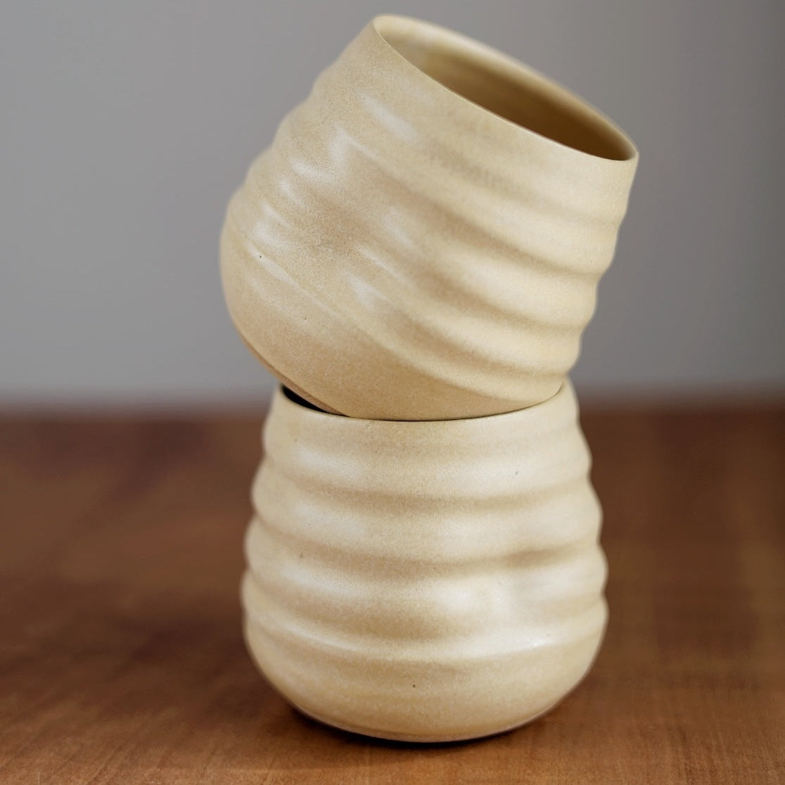 Two beige ceramic cups stacked on a wooden surface with a neutral background