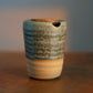 A handmade ceramic cup with a textured blue and green glaze on the upper half, a beige lower half, and a small hole near the rim, sitting on a wooden surface.
