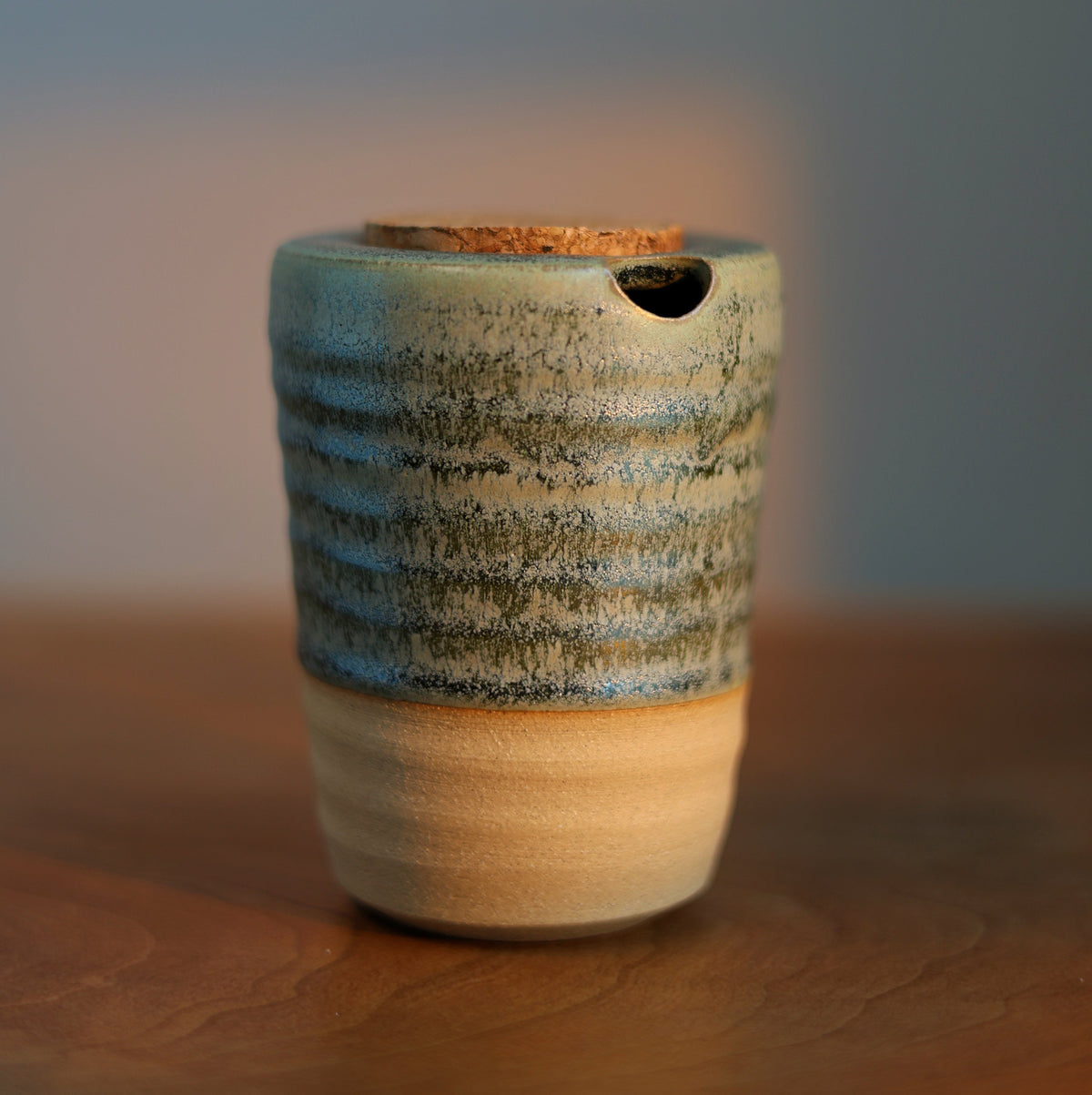 A handmade ceramic cup with a textured blue and green glaze on the upper half, a beige lower half, and a small hole near the rim, sitting on a wooden surface.