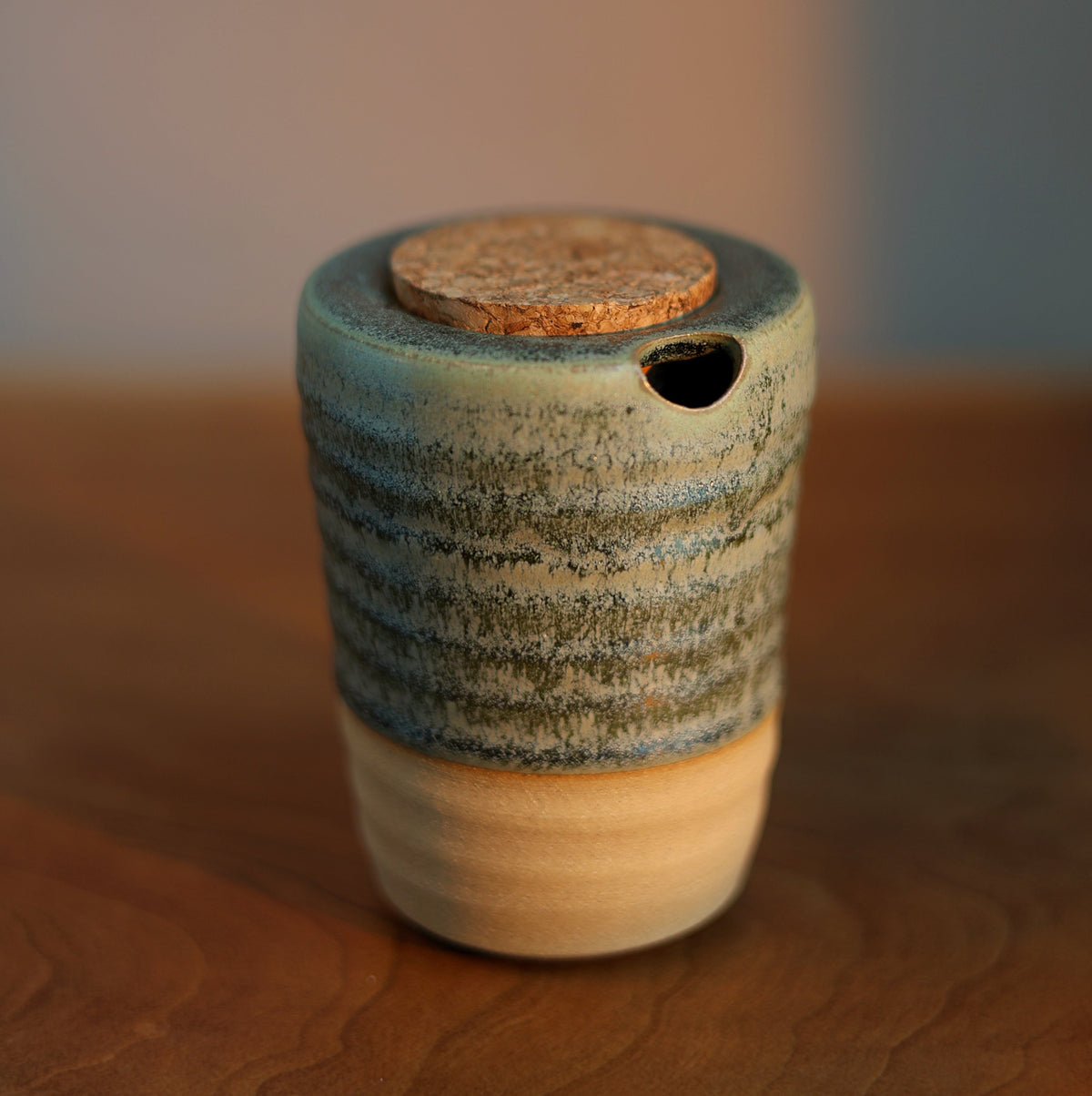 A small handmade ceramic container with a lid, featuring a textured blue and green glazed upper body, a natural beige clay base, and a cork stopper, set on a wooden surface with a blurred background.