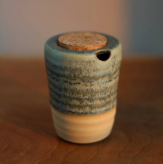 A small handmade ceramic container with a lid, featuring a textured blue and green glazed upper body, a natural beige clay base, and a cork stopper, set on a wooden surface with a blurred background.