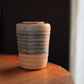 A small handmade ceramic cup with a textured, speckled glaze in green, beige, and brown tones, sitting on a wooden surface against a dark background.