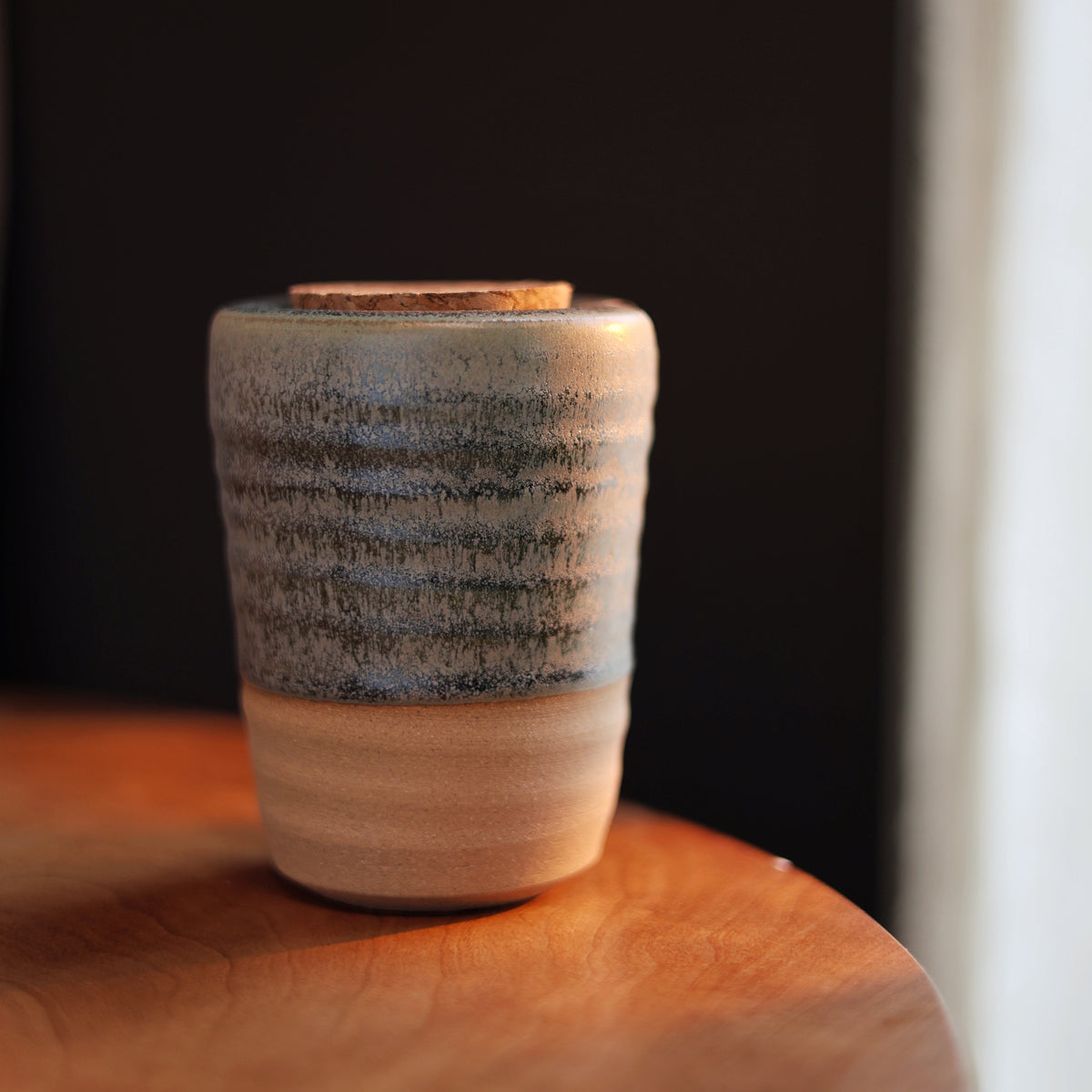 A small handmade ceramic cup with a textured, speckled glaze in green, beige, and brown tones, sitting on a wooden surface against a dark background.