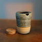 A small handmade ceramic cup with a textured blue-green and beige glaze sits on a wooden table next to a cork stopper, with a softly blurred background.