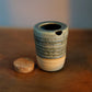 A small handmade ceramic vessel with a textured blue and beige glaze sits on a wooden surface next to a cork stopper, with a softly blurred blue background.