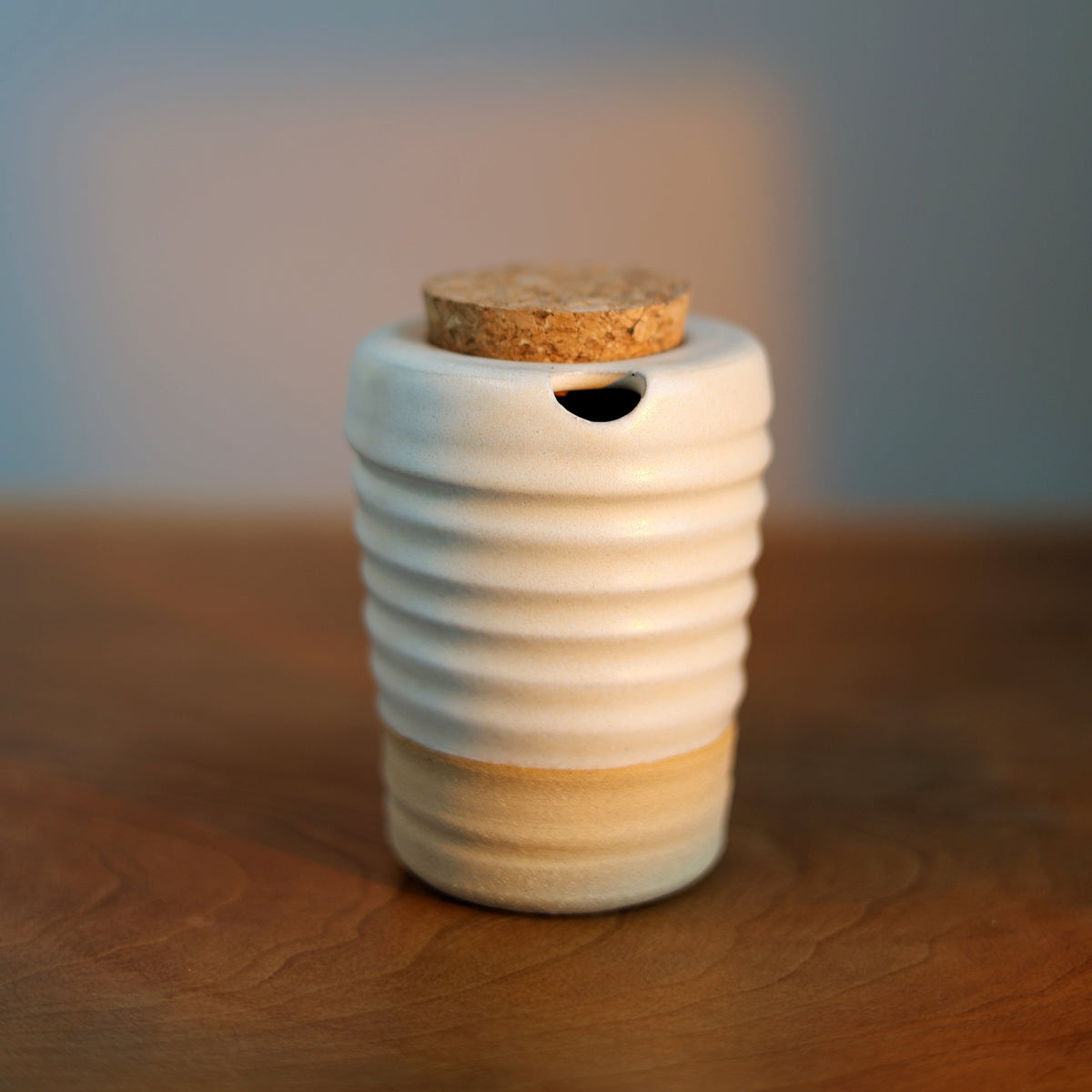 A small ceramic travel cup with a ribbed white body, a natural cork lid, and a light beige band near the base, sitting on a wooden surface against a softly blurred blue-gray background.
