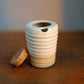 A small ribbed ceramic vessel with a white upper body and a warm beige lower band, shown on a wooden surface with a cork stopper beside it.