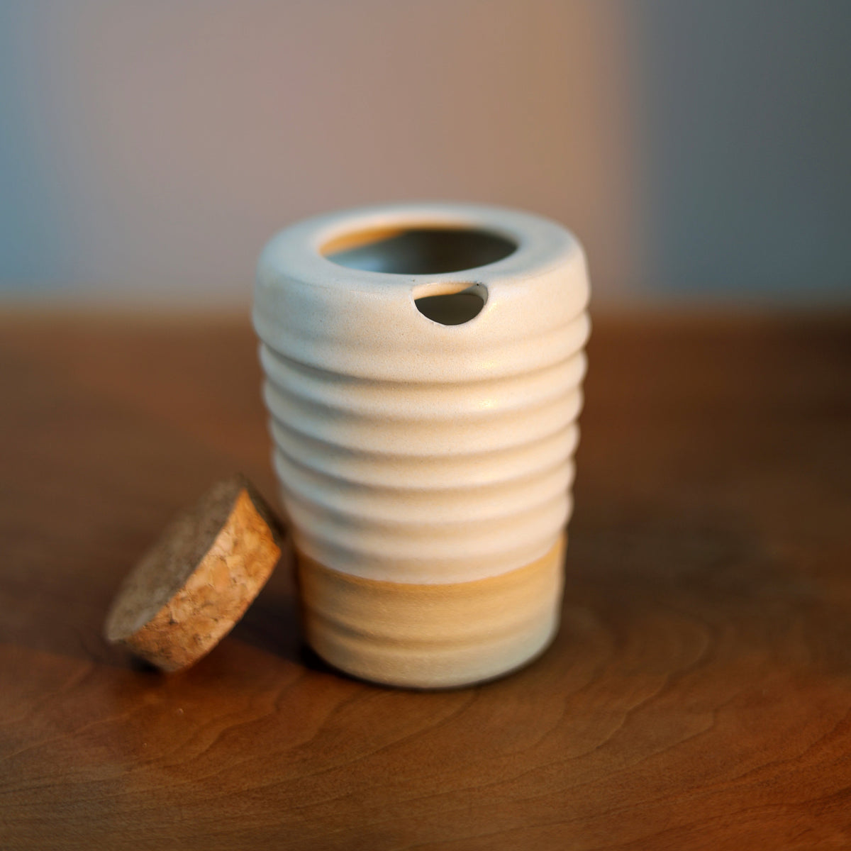 A small ribbed ceramic vessel with a white upper body and a warm beige lower band, shown on a wooden surface with a cork stopper beside it.