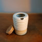 A ribbed ceramic mug with a white top half and a beige bottom half, sitting on a wooden table next to a cork stopper.