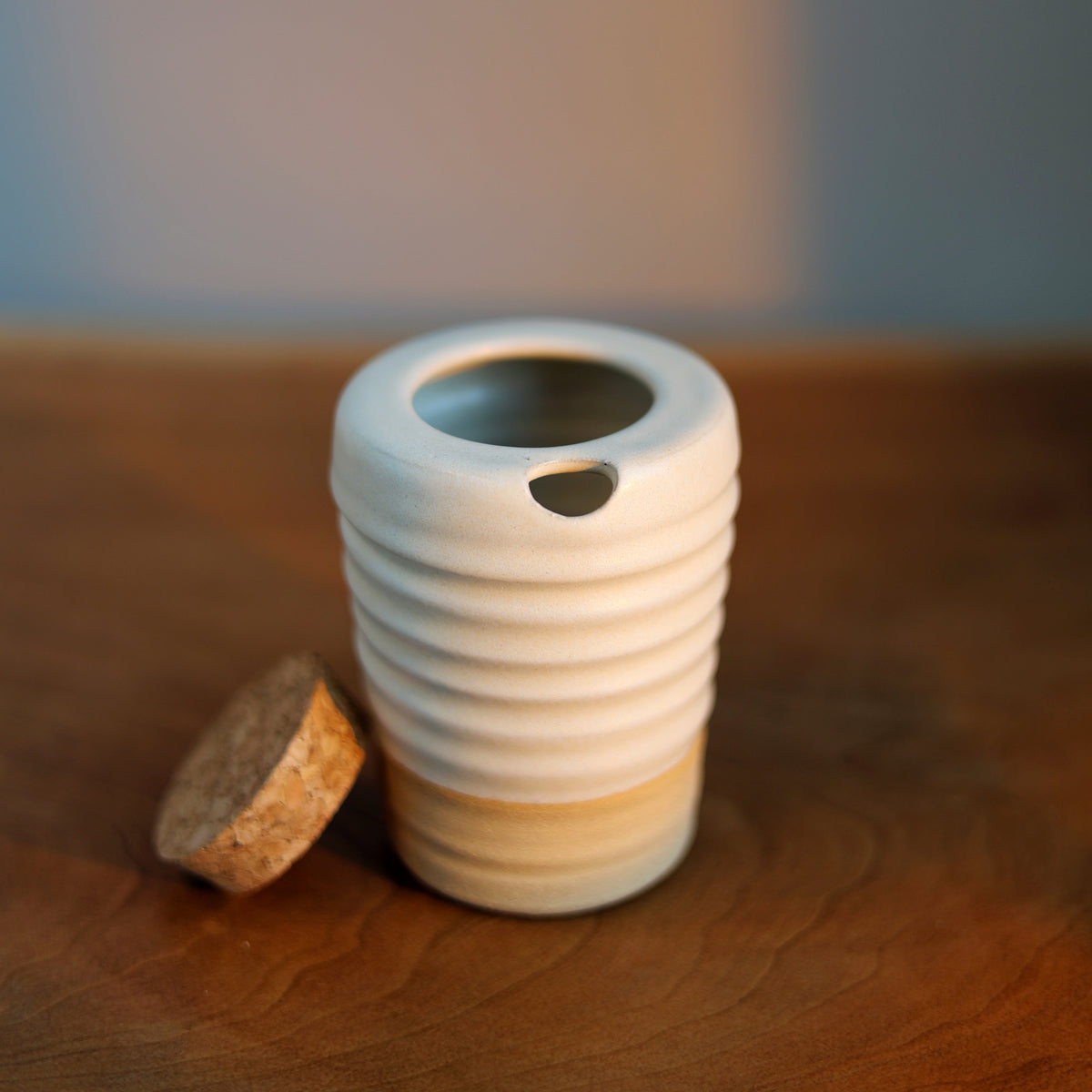 A ribbed ceramic mug with a white top half and a beige bottom half, sitting on a wooden table next to a cork stopper.