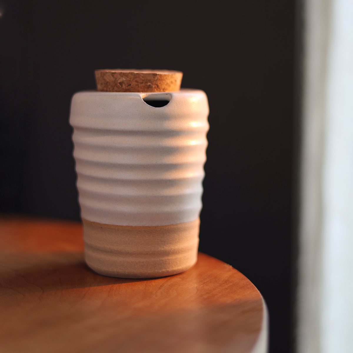 A small ceramic cup with a cork lid, featuring a white ribbed upper body and a natural beige base, sitting on a wooden surface against a dark background.