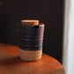 A small ceramic cup or container with a dark charcoal-black upper half and a natural light wood lower half, featuring a cork stopper and a small pouring spout, resting on a wooden surface against a dark background.