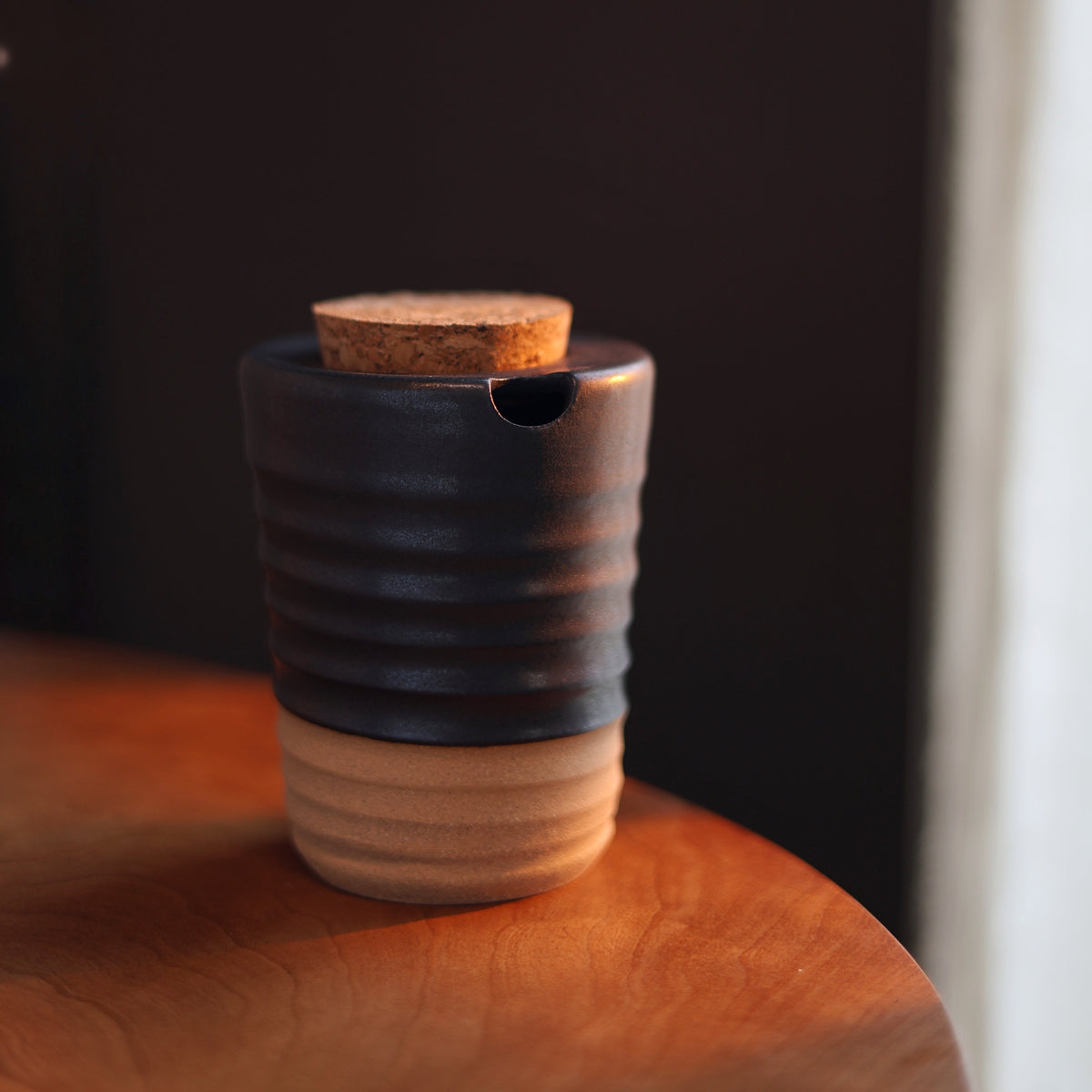 A small ceramic cup or container with a dark charcoal-black upper half and a natural light wood lower half, featuring a cork stopper and a small pouring spout, resting on a wooden surface against a dark background.