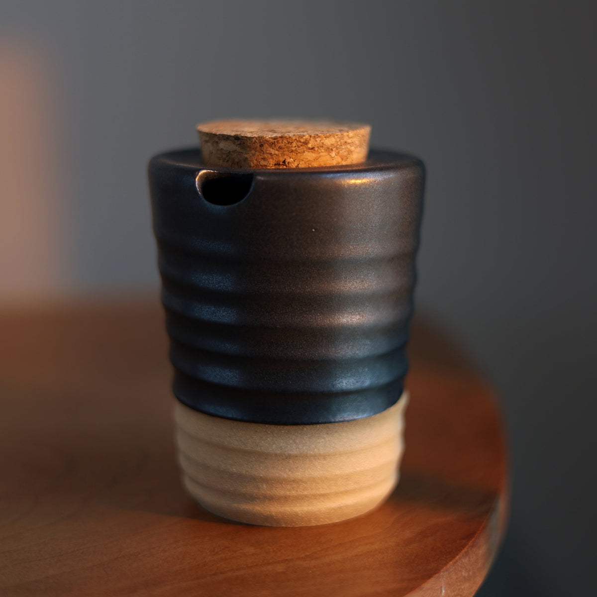 A small ceramic vessel with a dark charcoal ribbed body, a cork stopper, and a light natural wood base, resting on a wooden surface against a dark gray background.