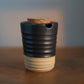 A small ceramic vessel with a dark charcoal-black upper half and a warm beige lower half, featuring a cork stopper and a small pouring spout, set on a wooden surface with a softly blurred background.