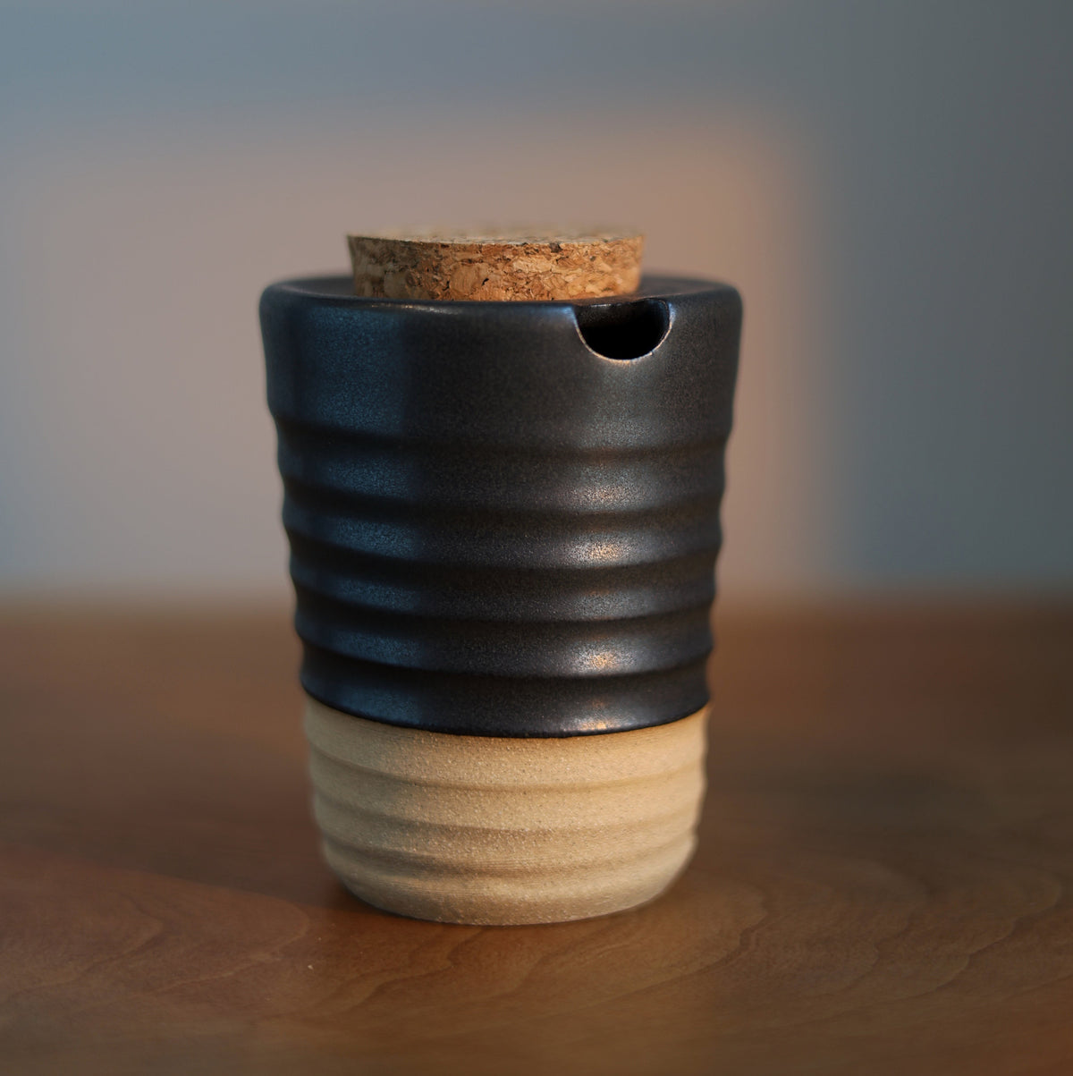 A small ceramic vessel with a dark charcoal-black upper half and a warm beige lower half, featuring a cork stopper and a small pouring spout, set on a wooden surface with a softly blurred background.