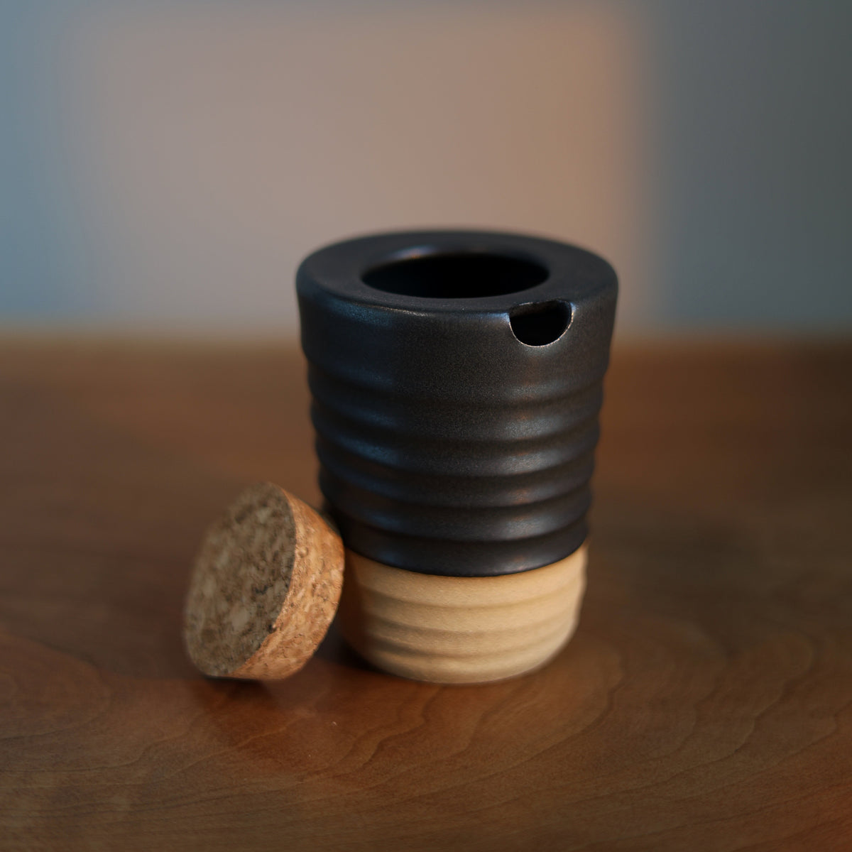 A small black ceramic vessel with a ribbed body and a small pouring spout, shown on a wooden surface with a cork stopper beside it. The background is softly blurred with warm light patches.