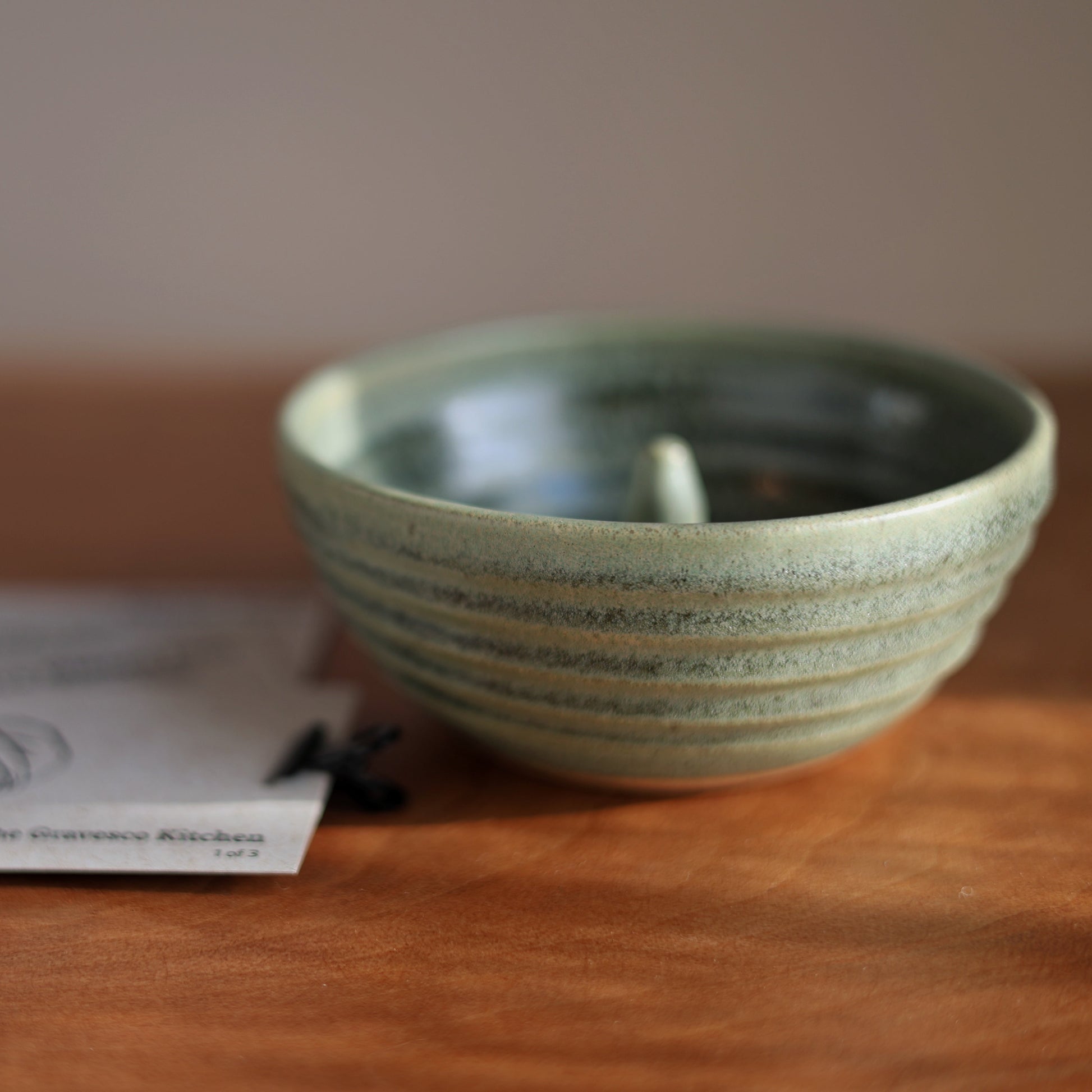 A small round ceramic apple baker with a speckled sage green glaze and horizontal ridges, sitting on a wooden table next to printed cards.